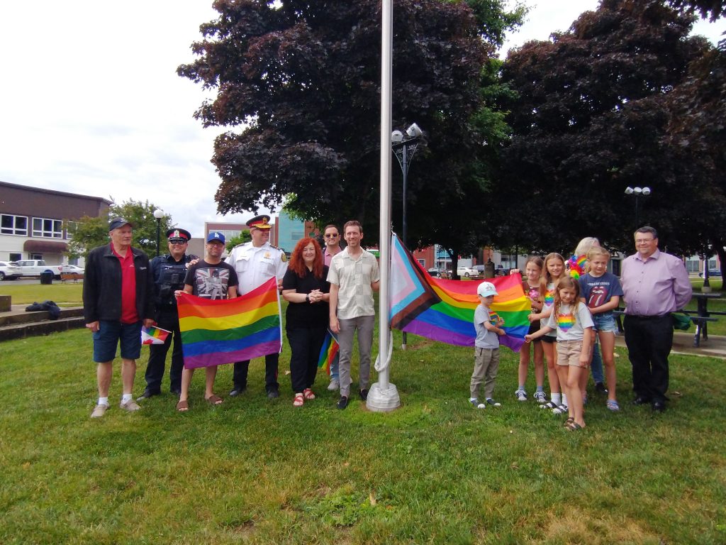 Un groupe, composé des officiels de la ville de Miramichi à gauche et d'un groupe de jeunes enfants à droite, posent de chaque côté d'une pôle à drapeaux avec celui de la fierté.