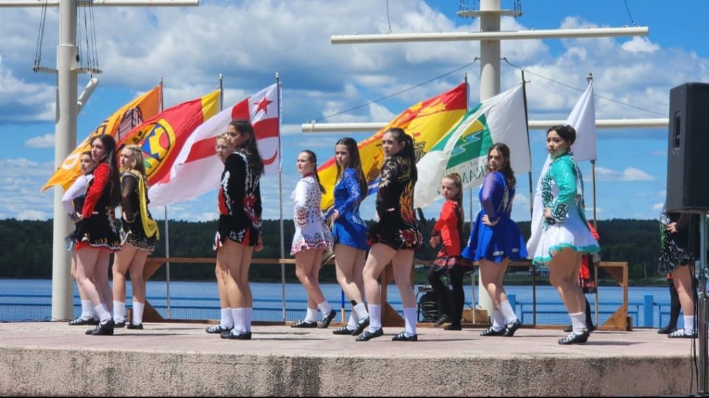 Un groupe de danse en pleine p^restation, en tenues irlandaises traditionnelles, devant plusieurs drapeaux flottant au vent, par journée ensoleillée.