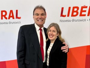 Sam Johnston et Susan Holt posent pour une photo, devant un fond aux couleurs du Parti libéral du Nouveau-Brunswick.