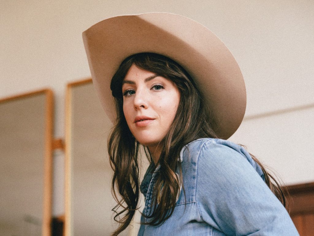 Assise sur une chaise vêtue de vêtements de jean et d'un chapeau de cowboy, Julie Aubé sourit à la caméra.