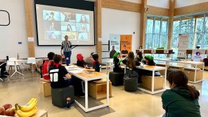 Plan large sur une vaste salle, avec une présentation devant des équipes de jeunes assis à des tables en groupe.