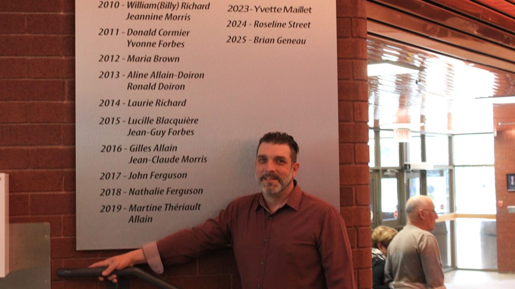 Photographie de Brian Geneau devant la plaque commémorative des pionniers du Carrefour Beausoleil.