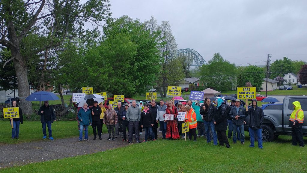 Photographie d'un groupe de personnes, pancartes à la main, avec l'arche principale du pont Centenaire en arrière-plan.