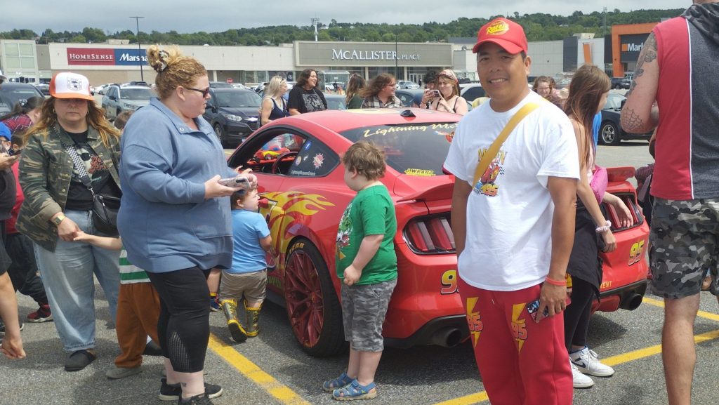 Gary Papaya, habillé en rouge et blanc avec du merch Cars pose devant sa voiture, une Mustang rouge avec des stickers de Flash McQueen