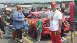 Gary Papaya, habillé en rouge et blanc avec du merch Cars pose devant sa voiture, une Mustang rouge avec des stickers de Flash McQueen