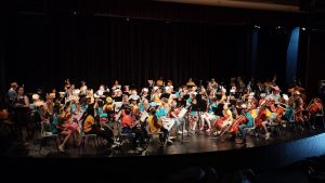 125 enfant sur scene habillés de t-shirt colorés et avec chacun leur instrument
