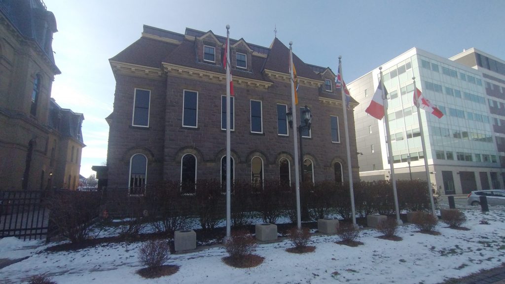 Photographie de l'aile ouest de l'Assemblée législative du Nouveau-Brunswick, devant lequel se trouve cinq drapeaux.